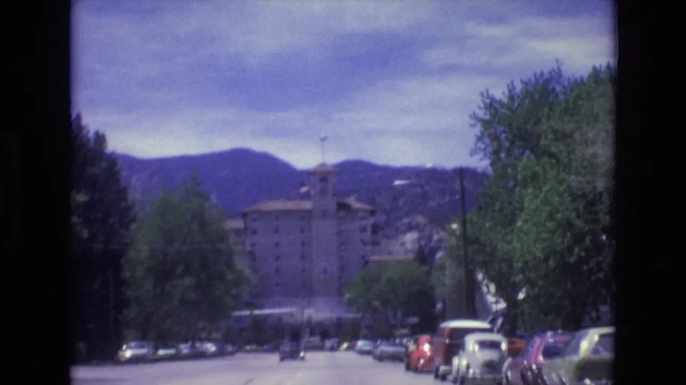 1973 Colorado Springs frame from a moving vehicle, with classic cars along the road, a prominent building ahead, and mountains across the horizon.
