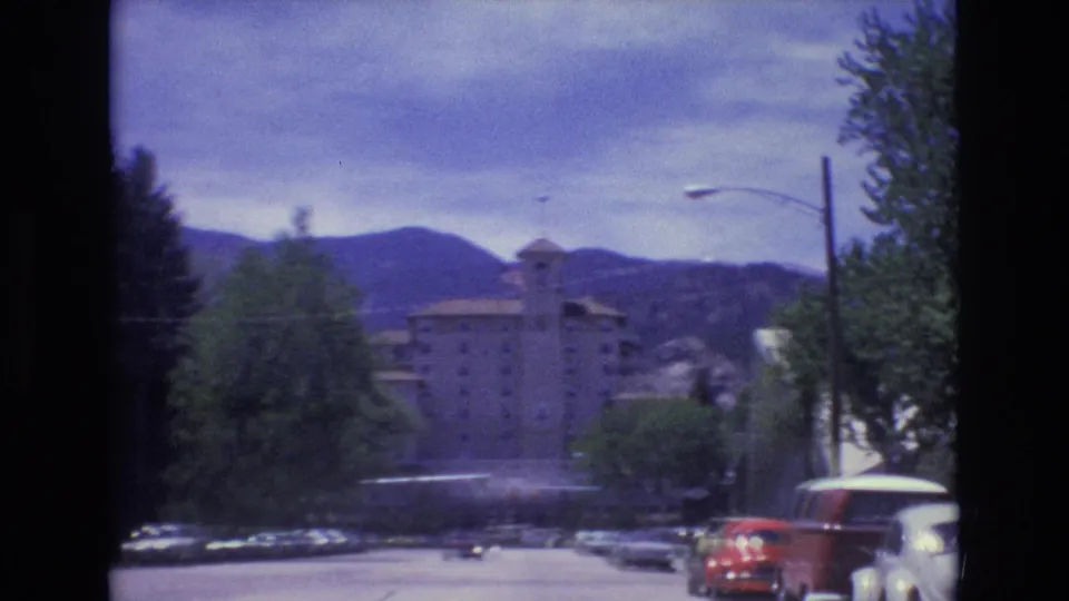 Motion-blurred 1973 Colorado Springs street frame showing a large building ahead, roadside trees, parked cars, a streetlight, and mountains beyond.