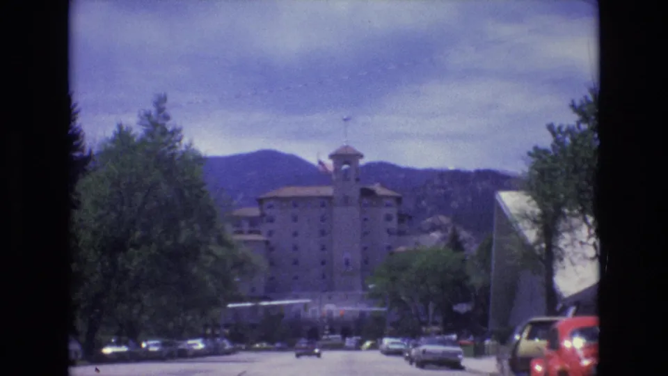 Late frame of the 1973 Colorado Springs road sequence, showing cars along the street, trees, the distant towered building, mountains, and an angular building edge at right.