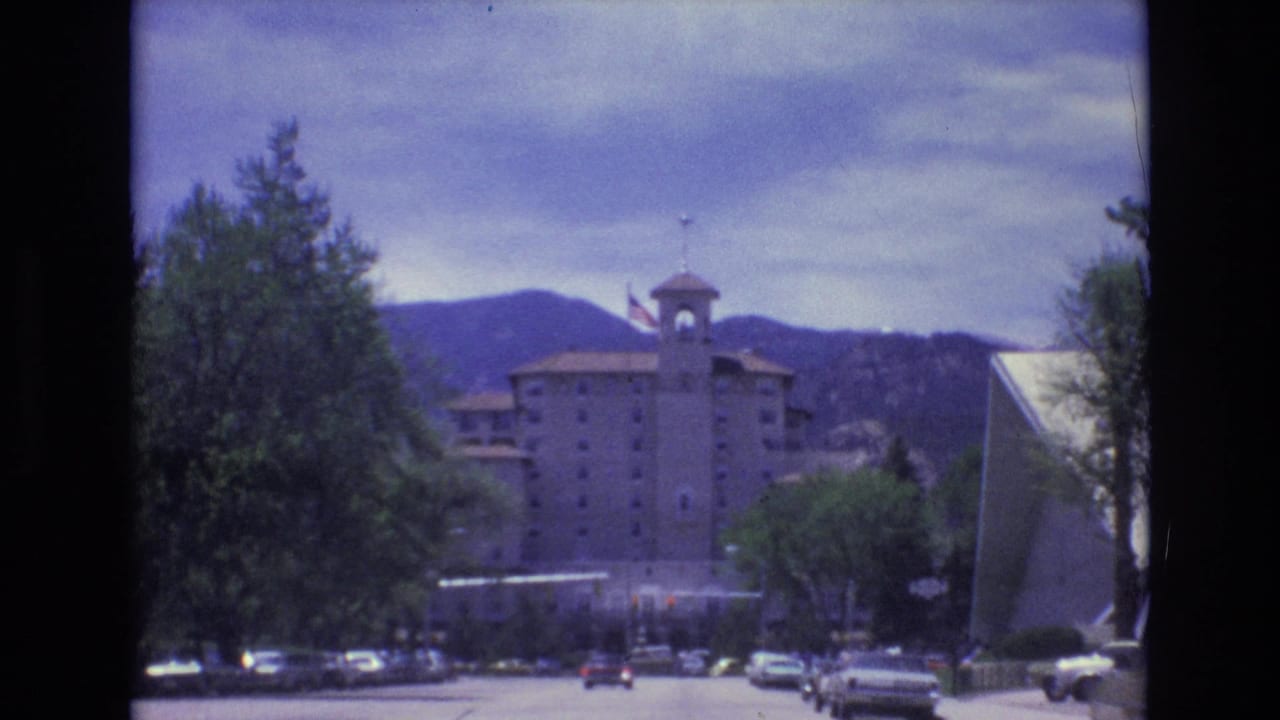 1973 Colorado Springs street view showing a towered hotel-like building with an American flag, parked and moving cars, roadside trees, and mountains in the background.