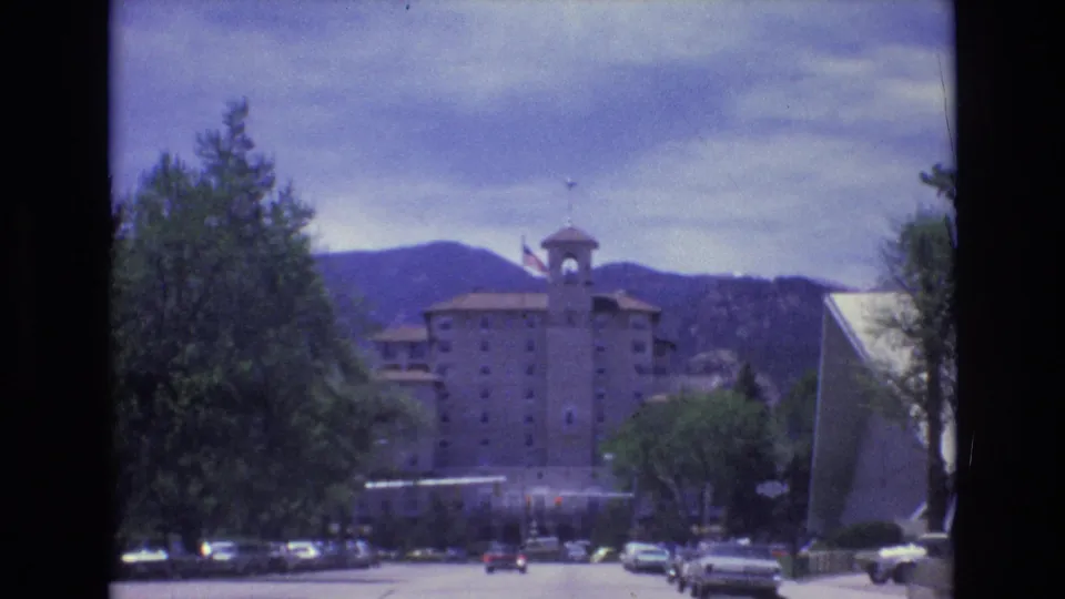 1973 Colorado Springs street view showing a towered hotel-like building with an American flag, parked and moving cars, roadside trees, and mountains in the background.