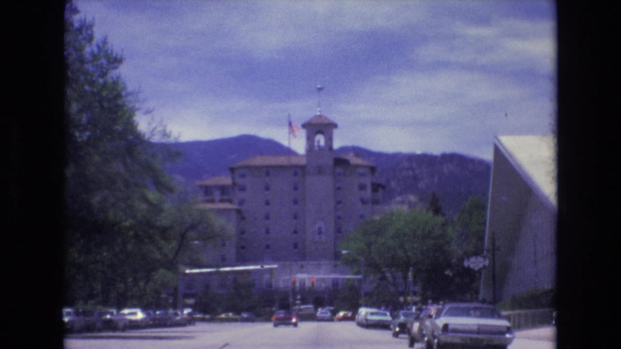 1973 Colorado Springs frame of a towered building and flag beyond a street with classic cars, leafy trees, and low mountains in the distance.