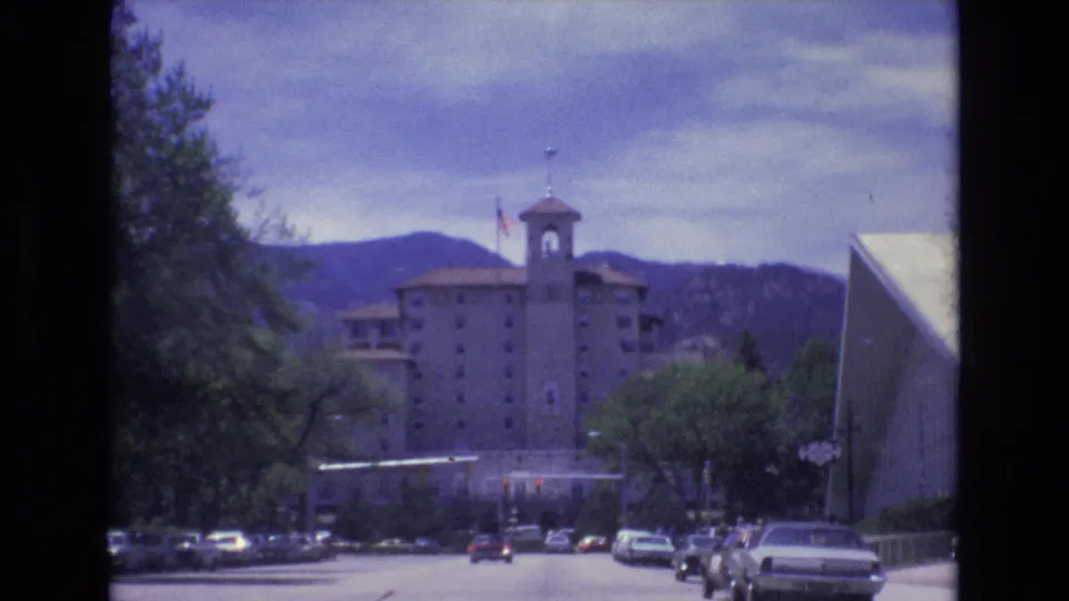 1973 Colorado Springs frame of a towered building and flag beyond a street with classic cars, leafy trees, and low mountains in the distance.
