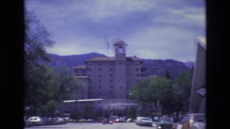 Soft 1973 8mm view in Colorado Springs with the towered building centered behind trees, cars along the street, a flagpole, and mountains beyond.