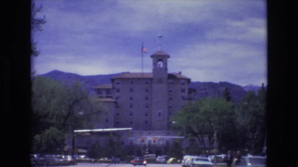 1973 Colorado Springs view with the towered building slightly closer in frame, an American flag overhead, cars at street level, trees, and mountains behind.