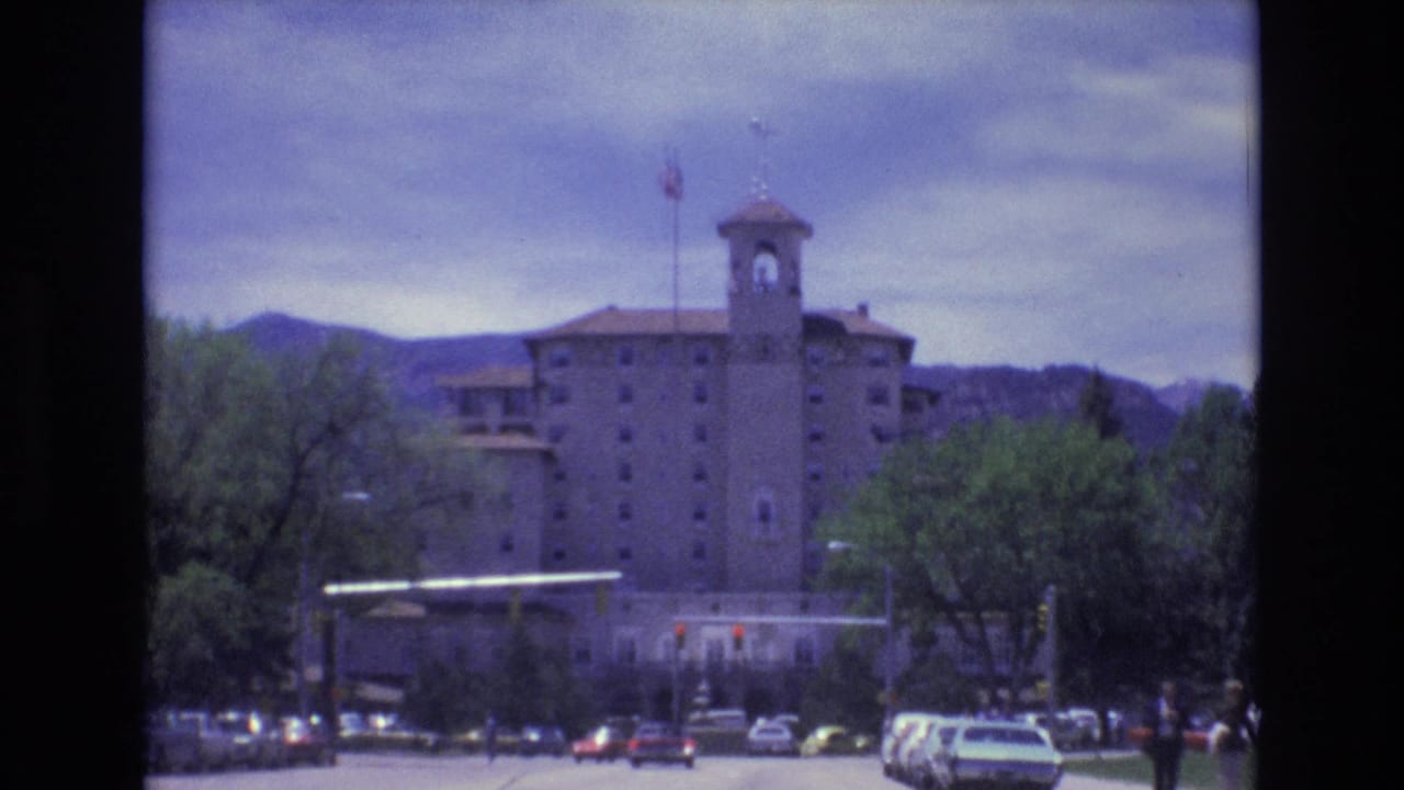 1973 Colorado Springs street view with a towered multi-story building, American flag, cars near a red light, trees, and low mountains under cloudy sky.