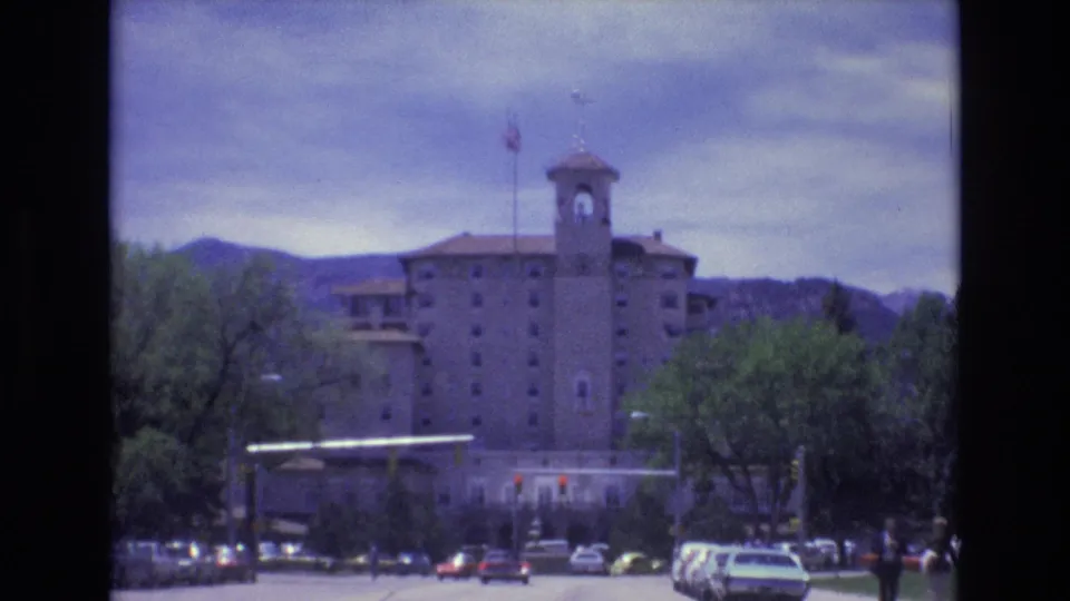 1973 Colorado Springs street view with a towered multi-story building, American flag, cars near a red light, trees, and low mountains under cloudy sky.