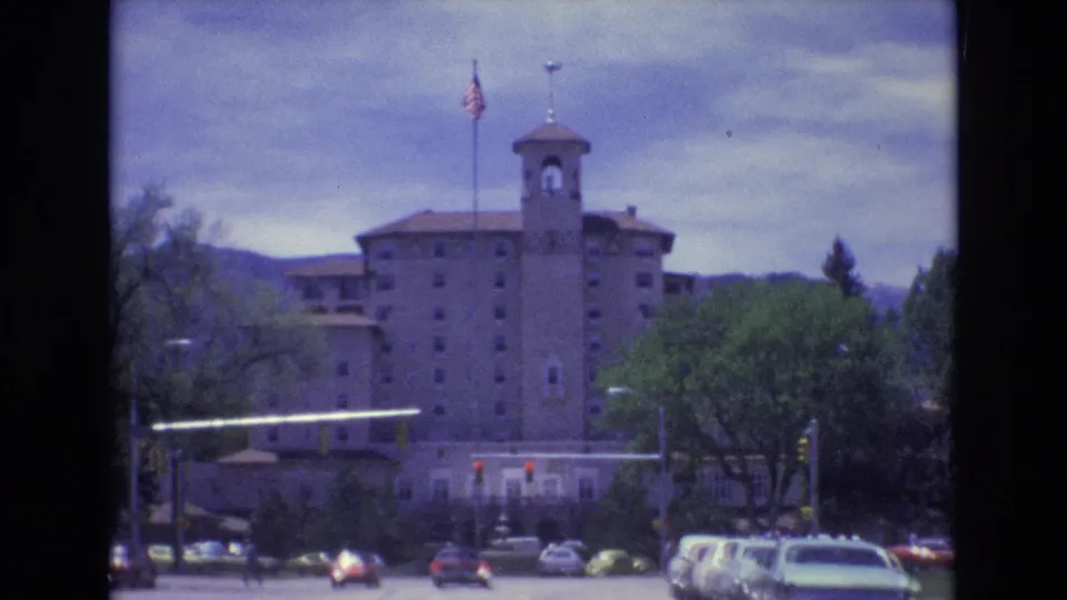 Colorado Springs street scene from 1973 with period cars, pedestrians, red traffic signals, trees, and a towered building set against distant mountains.