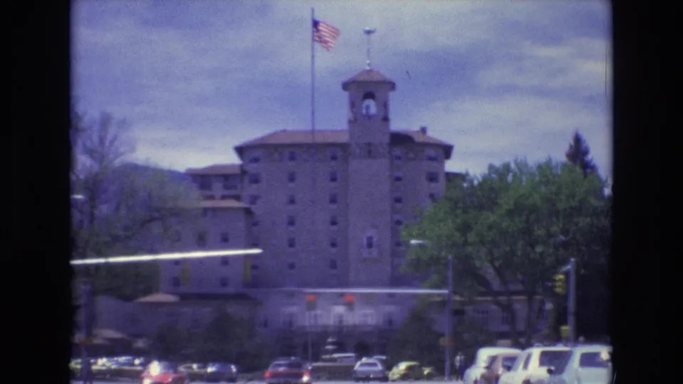 Closer 1973 Colorado Springs view of a tall towered building, American flag, red traffic signals, cars, trees, and hazy mountains in the background.