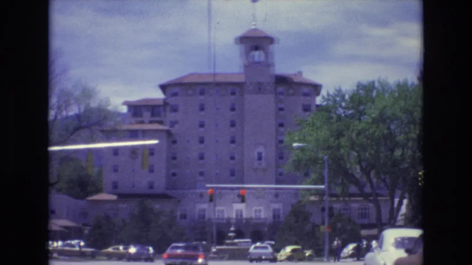 1973 Colorado Springs home-movie frame showing a towered building, traffic stopped at red lights, nearby pedestrians, trees, parked cars, and cloudy mountain backdrop.