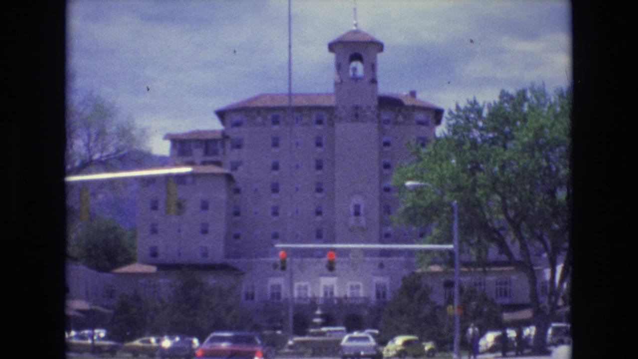 Colorado Springs 1973 home-movie frame showing a large multi-story building with a bell tower, cars at an intersection, and trees under a cloudy sky.