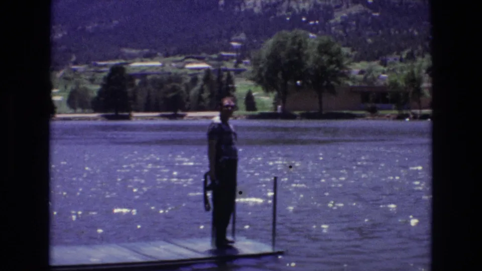 A man stands in profile on a wooden dock by a bright lake in 1973 Colorado Springs home-movie footage, with trees, buildings, and mountains across the water.