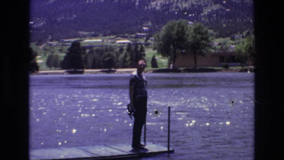 The man is still standing on the dock in the 1973 Colorado Springs lake scene, framed against sparkling water, shoreline trees, and distant mountains.