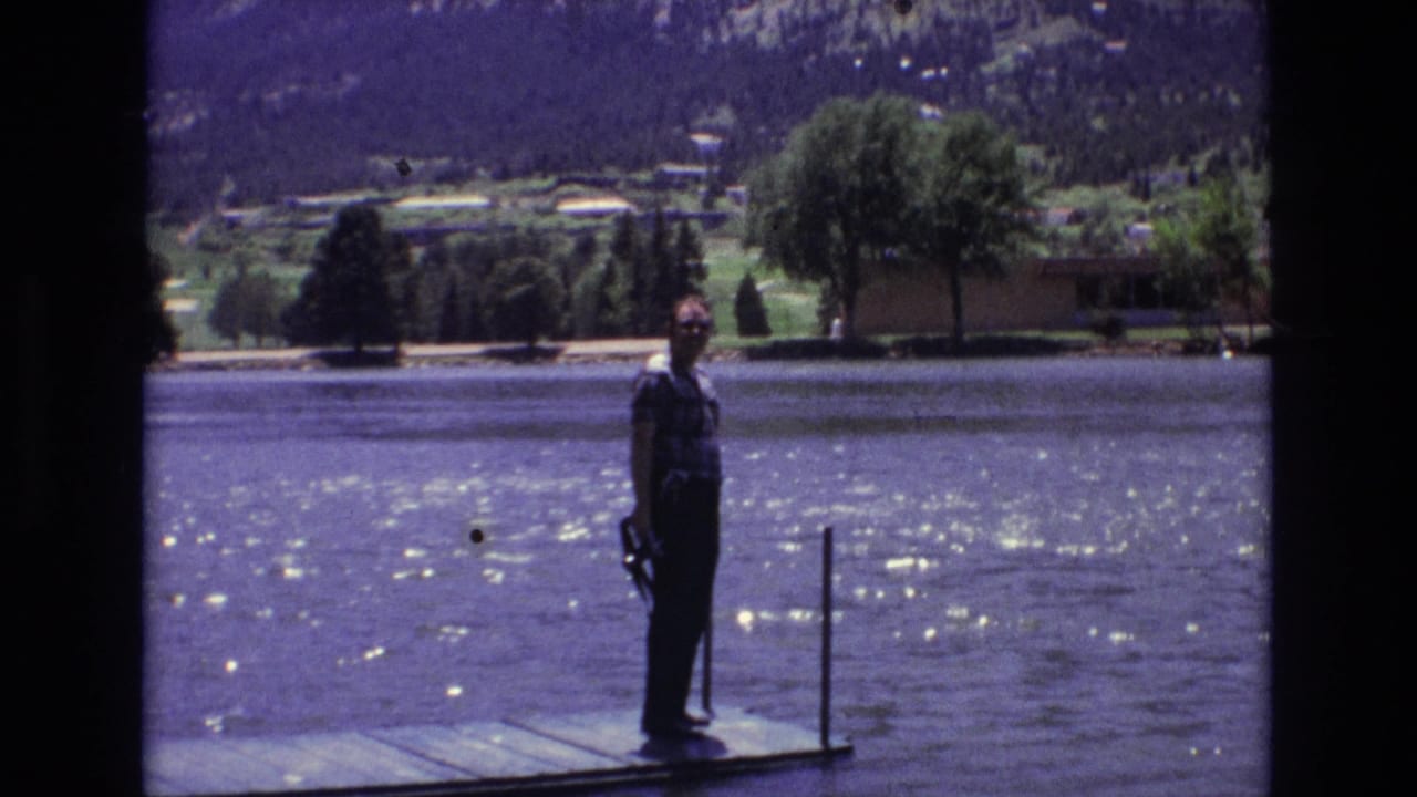 1973 Colorado Springs home-movie frame showing a man in a plaid shirt standing on a wooden dock beside sparkling lake water with trees, distant houses, and mountains behind him.