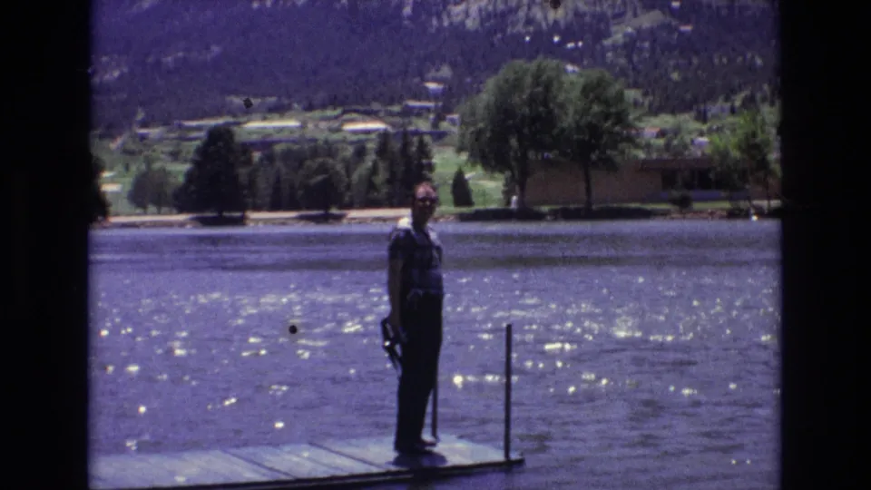 1973 Colorado Springs home-movie frame showing a man in a plaid shirt standing on a wooden dock beside sparkling lake water with trees, distant houses, and mountains behind him.