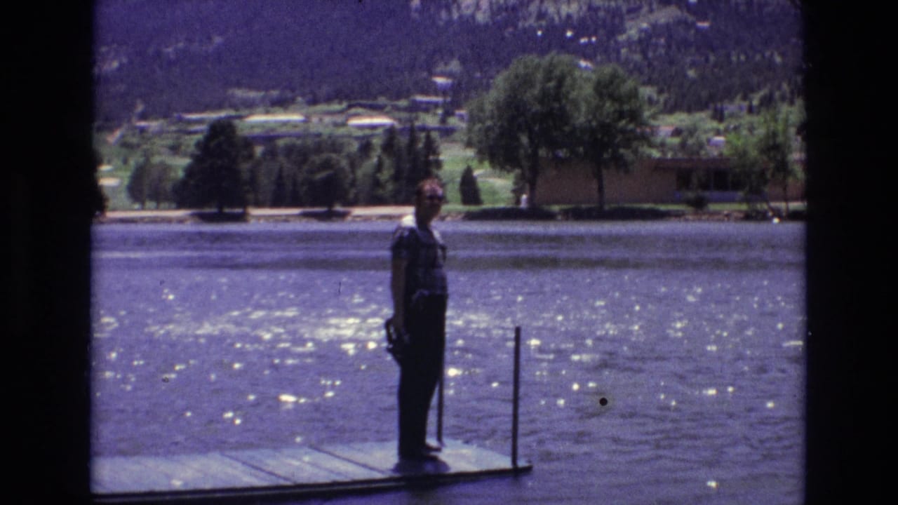 1973 Colorado Springs lakeside frame with a man standing on the dock facing the camera, water glinting in sunlight, and trees and mountain slopes across the lake.