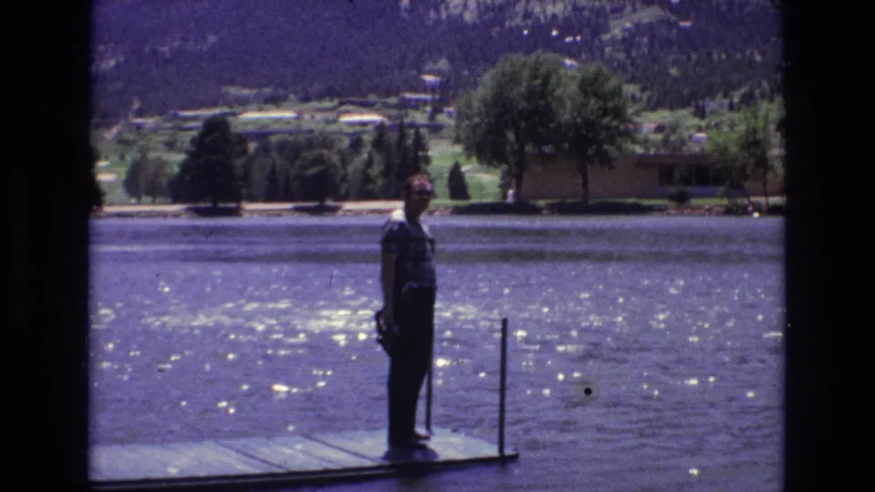 1973 Colorado Springs lakeside frame with a man standing on the dock facing the camera, water glinting in sunlight, and trees and mountain slopes across the lake.