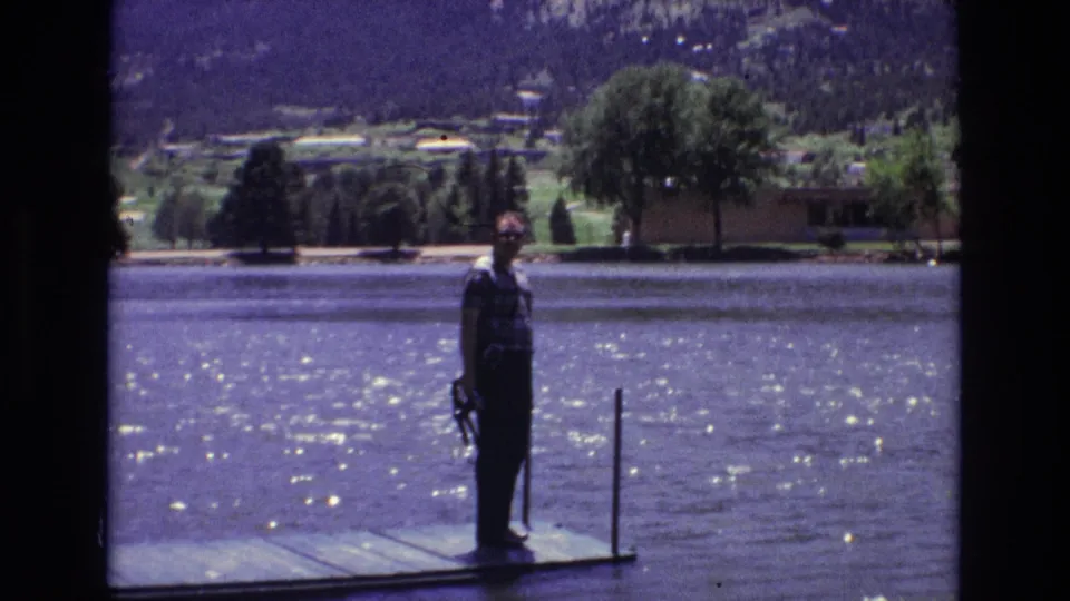1973 Colorado Springs home-movie still of a man standing near a dock post on a lakeside platform, with rippled water, trees, houses, and mountain terrain in the distance.