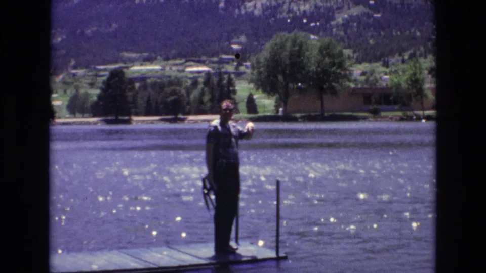 1973 Colorado Springs frame showing the man raising one hand in a wave from the dock, with sparkling lake water, shoreline trees, distant houses, and mountains visible behind him.