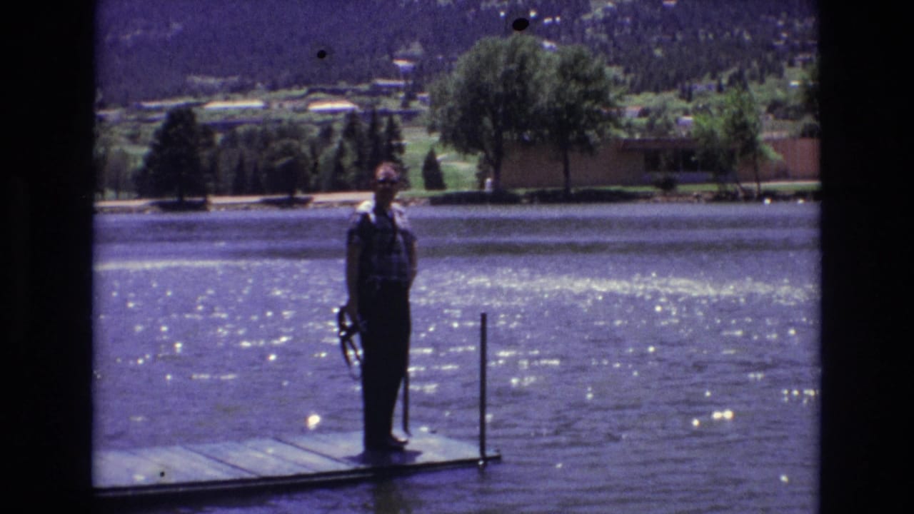 Man with fishing rod standing on a dock over bright lake water.