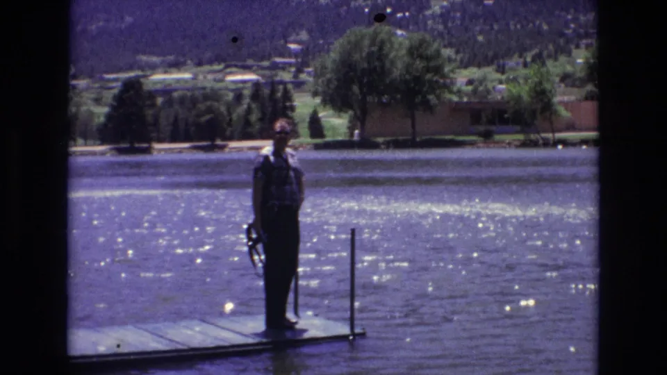 The 1973 Colorado Springs home-movie frame shows a man on a dock with a fishing rod, lake glare, trees, and a mountain backdrop.
