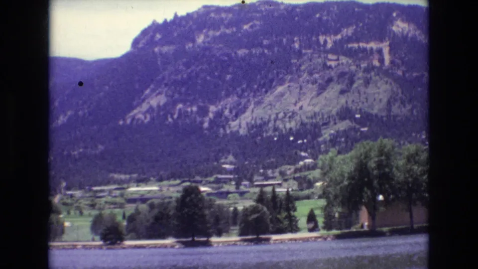 A Colorado Springs lakeside view shows rippled water, shoreline trees, low structures, and a forested mountain slope in 1970s 8mm color footage.