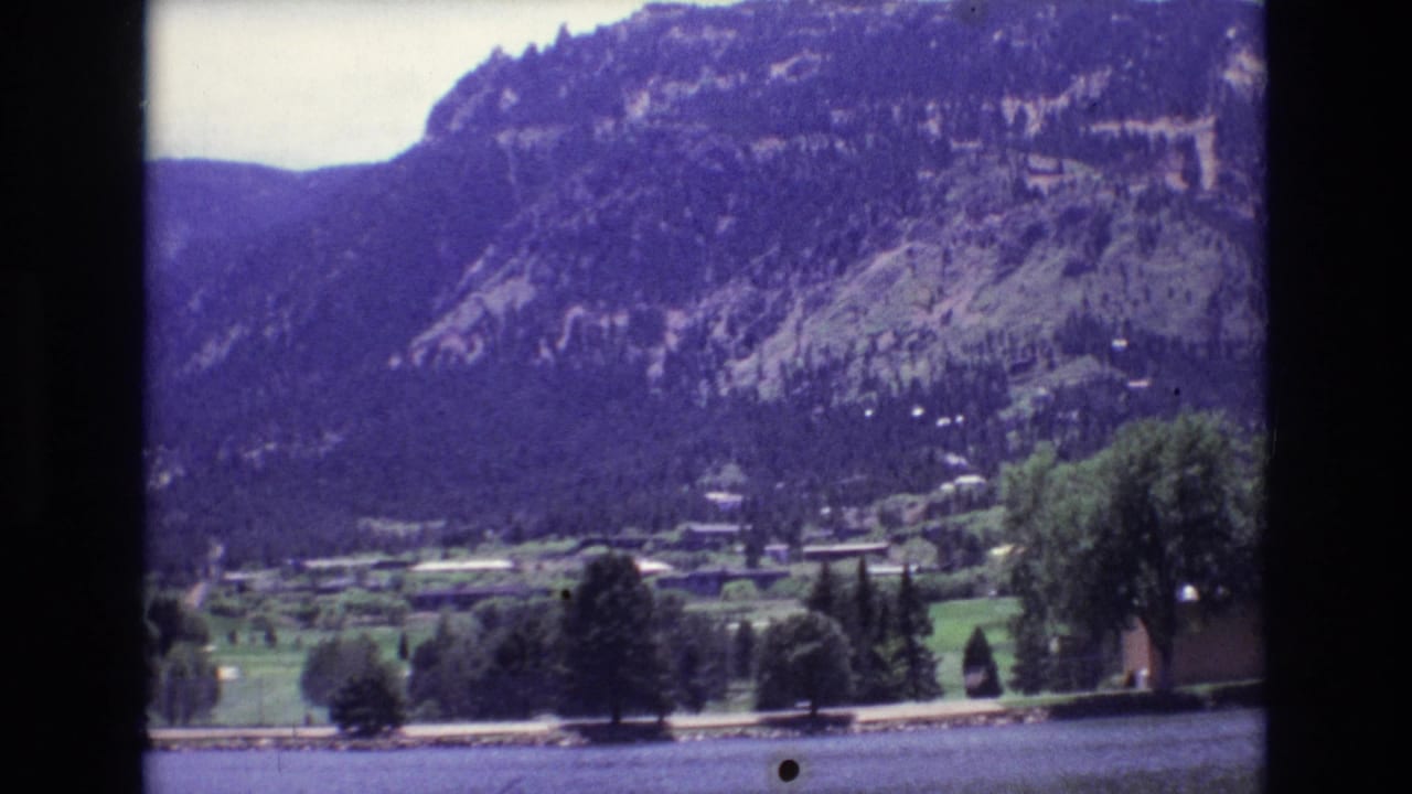 Colorado Springs 8mm frame with a calm water edge, valley trees, scattered buildings, and a broad forested mountain rising behind them.