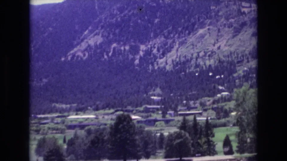 Zoomed 1973 home-movie frame showing trees, open green land, distant buildings, and a rocky forested mountain backdrop in Colorado Springs.