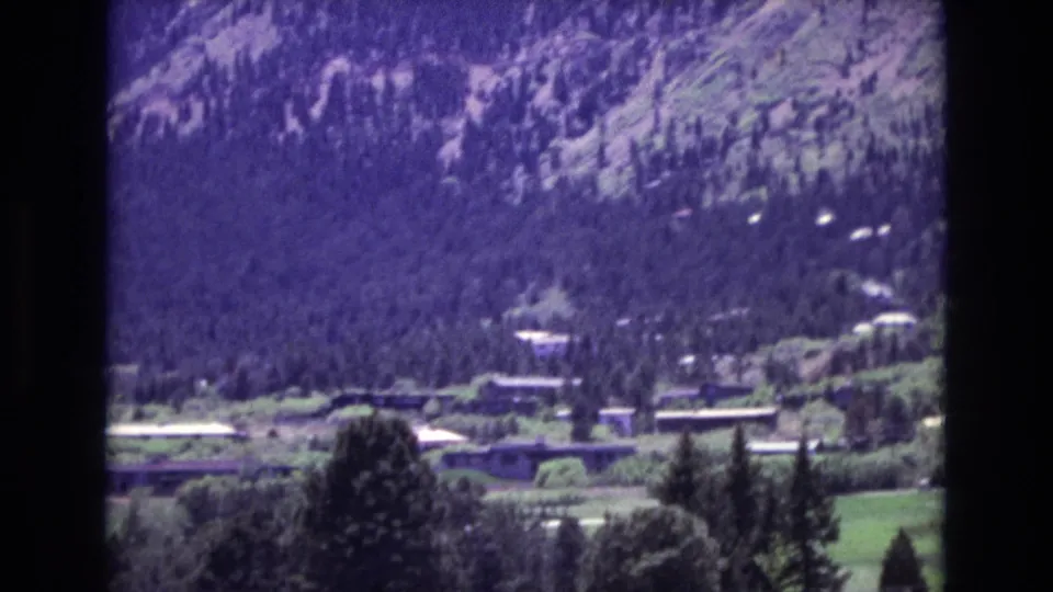 1973 8mm frame from Colorado Springs showing a closer valley view with trees, scattered buildings, road lines, and a forested mountain slope behind.