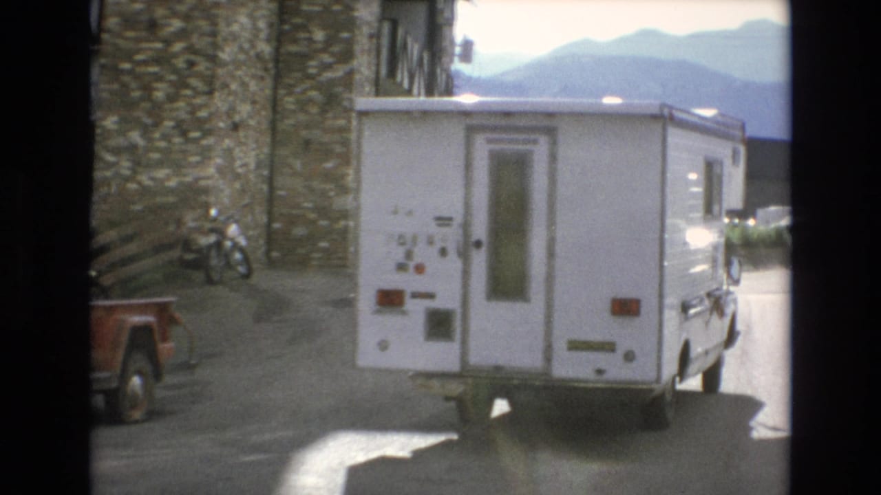 1973 Colorado Springs home-movie frame showing a white RV close to the camera beside a stone-faced resort building, with distant mountains under bright sky.