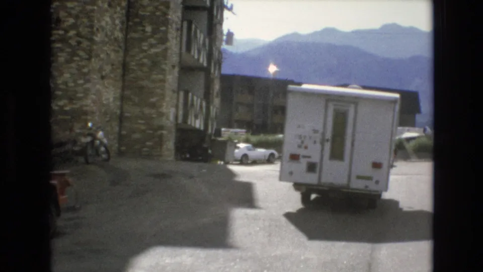 1973 Colorado Springs frame with the white recreational vehicle moving away through a paved mountain resort parking area, with lodging buildings and parked cars visible.