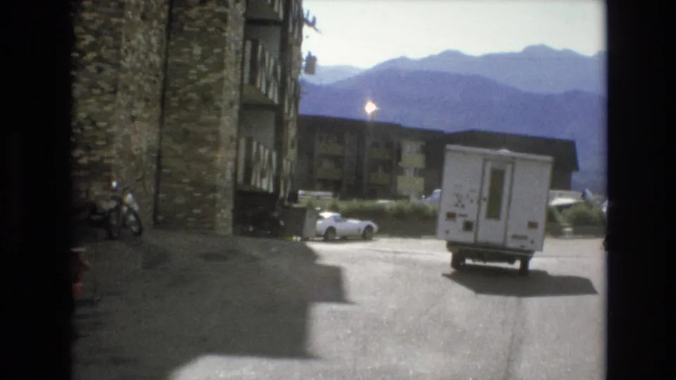 1973 Colorado Springs home-movie frame showing the RV angled away from the camera in a bright paved resort area, with parked cars, lodging, and layered mountains behind it.