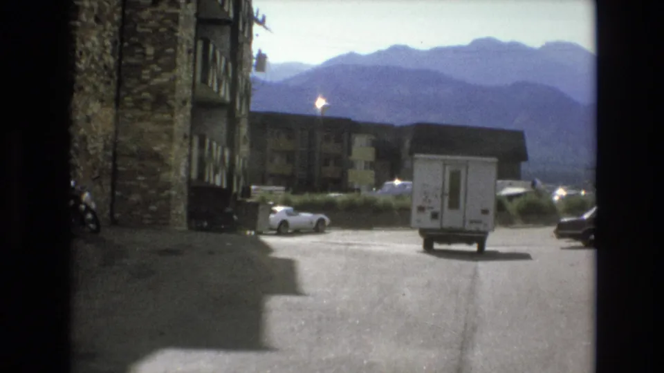1973 Colorado Springs frame showing the white RV farther away in the resort parking area, framed by low-rise buildings, parked cars, and a hazy mountain backdrop.