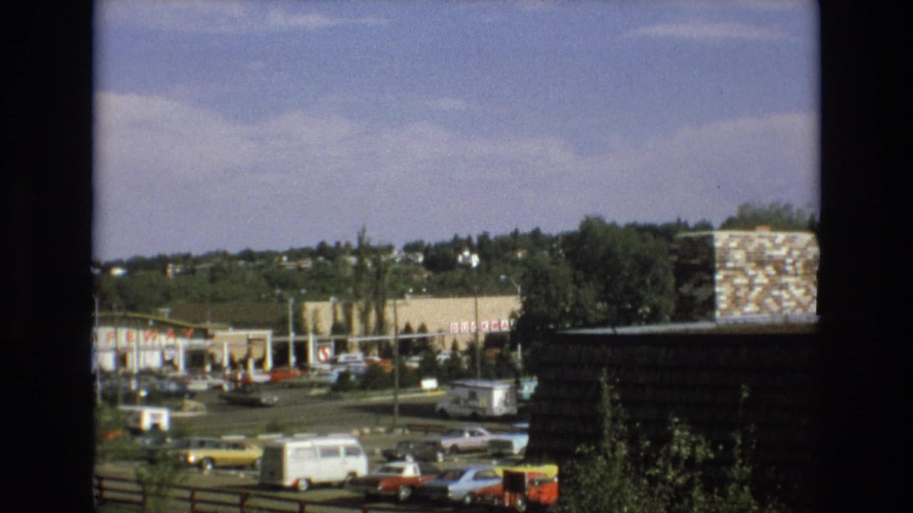 1973 Colorado Springs commercial area viewed from behind a dark window frame, with a surface parking lot, classic cars, a white van, storefronts, and a Safeway sign visible in the distance.