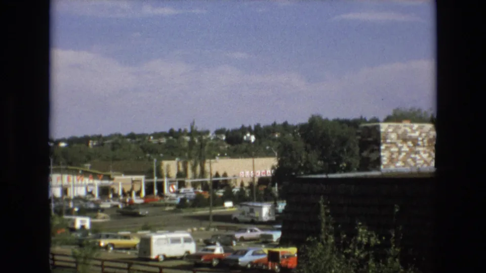 1973 Colorado Springs commercial area viewed from behind a dark window frame, with a surface parking lot, classic cars, a white van, storefronts, and a Safeway sign visible in the distance.