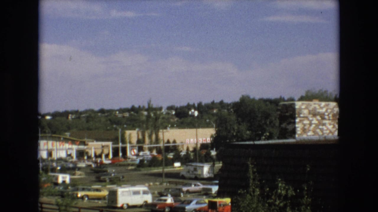 Commercial parking lot with parked cars, a white van, distant storefronts, trees, and a dark foreground border.