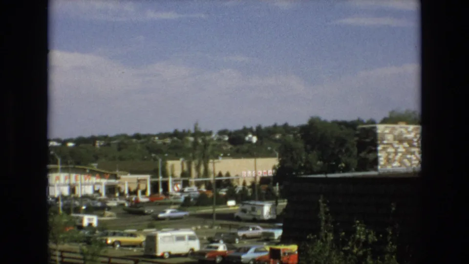 1973 home-movie frame of a Colorado Springs retail parking area with a white van, yellow sedan, multiple classic cars, storefronts including Safeway signage, and a distant tree-lined background.