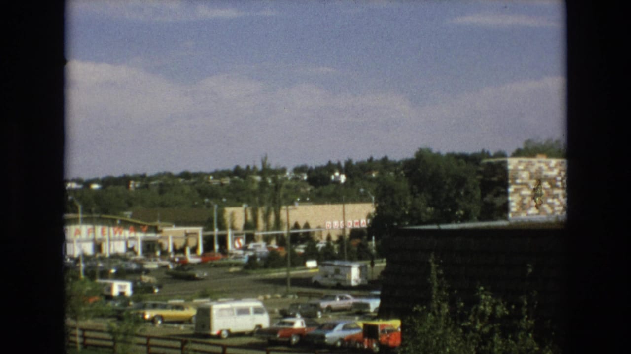 1973 Colorado Springs home-movie frame showing a shopping center parking lot with cars, a white van, Safeway signage, trees, and hillside residences in the background.