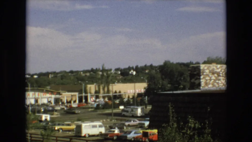1973 Colorado Springs 8mm view across a shopping center parking lot with a white van, yellow car, storefront signage, trees, and hillside homes.