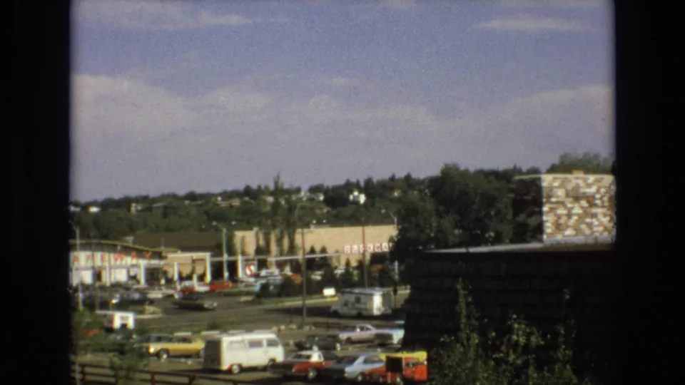 1973 Colorado Springs home-movie frame showing cars in a shopping center parking lot, Safeway signage, a white van, trees, and hillside housing beyond.