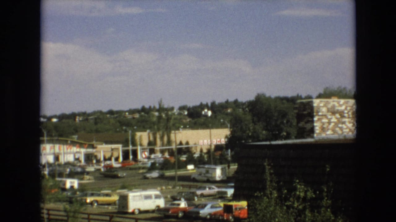 1973 Colorado Springs home-movie frame showing a sunlit commercial parking area with period cars, a white van, a grocery storefront, trees, distant hillside homes, and a dark foreground window edge.