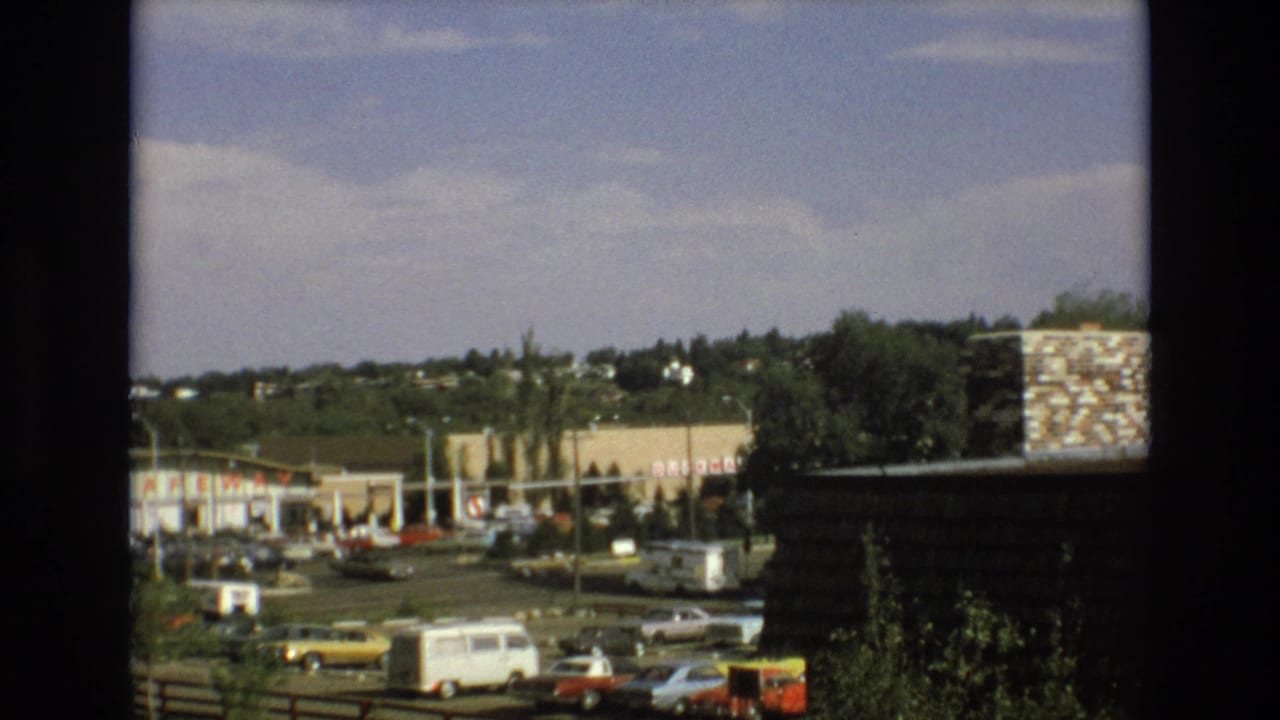Commercial parking lot with classic cars, a white van, storefront signs, trees, hillside houses, sky, and dark window framing.