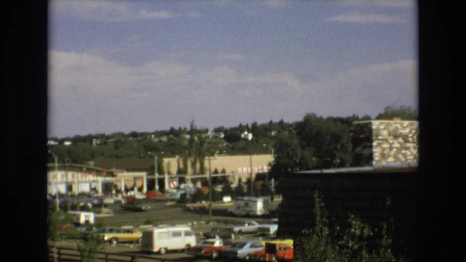 1973 Colorado Springs frame looking past a dark window edge toward a busy roadside shopping area with classic cars, a white van, visible storefront branding, trees, and homes on a hillside.