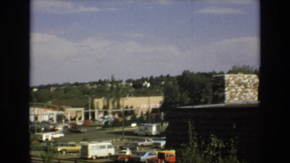 1973 Colorado Springs home-movie still with a slightly lower handheld view of a commercial parking lot, period vehicles, a white van, storefronts, trees, hillside houses, and a broad sky.