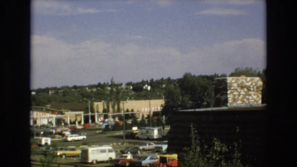 1973 Colorado Springs frame showing a roadside commercial district from behind dark window framing, with 1970s cars, a white van, storefronts, trees, and houses visible on the hillside beyond.