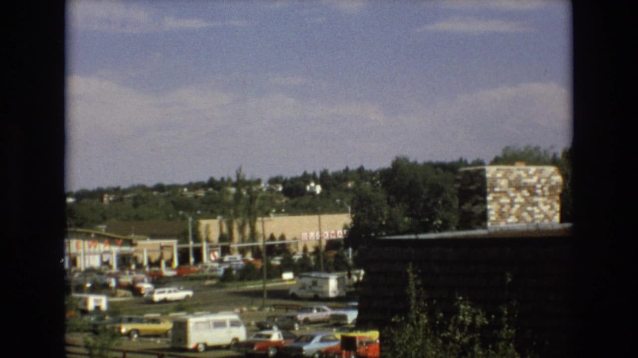 1973 Colorado Springs view of a suburban commercial area with Safeway signage, parked cars, a white van, trees, and a stone chimney in the foreground.