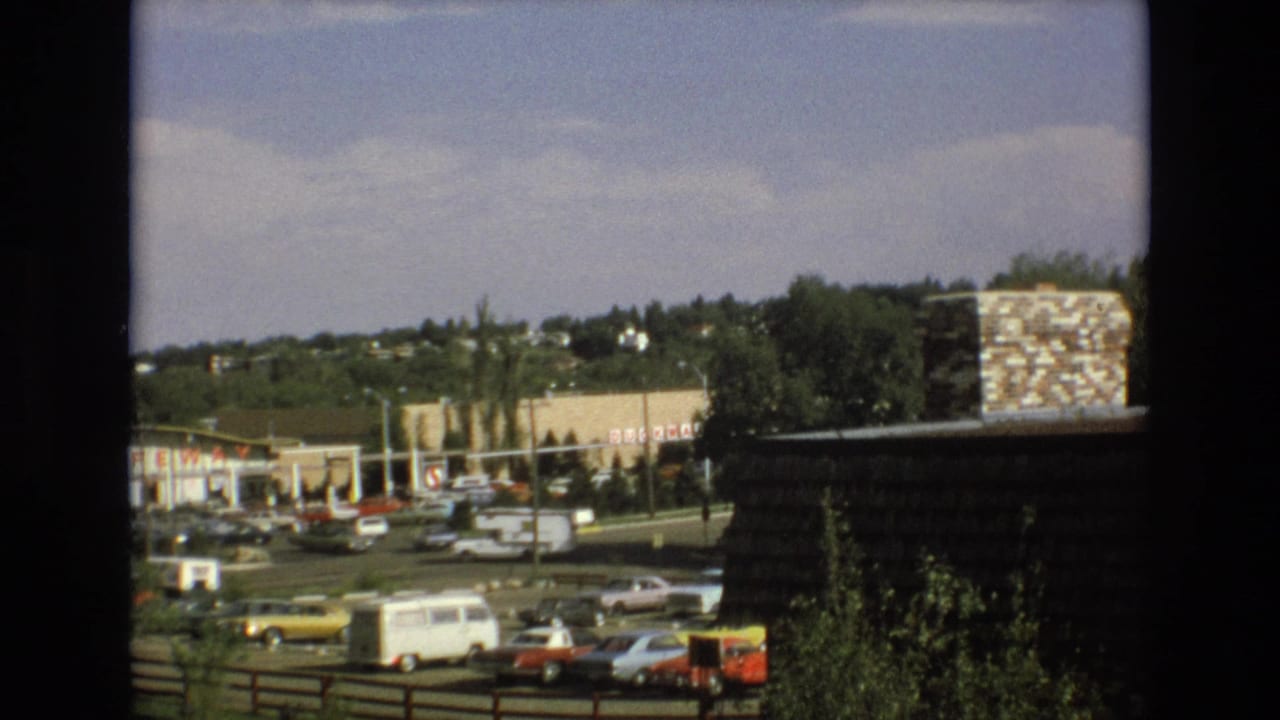 1973 Colorado Springs commercial strip with Safeway branding, a busy parking lot, a white van, period sedans, trees, and a stone chimney at right.