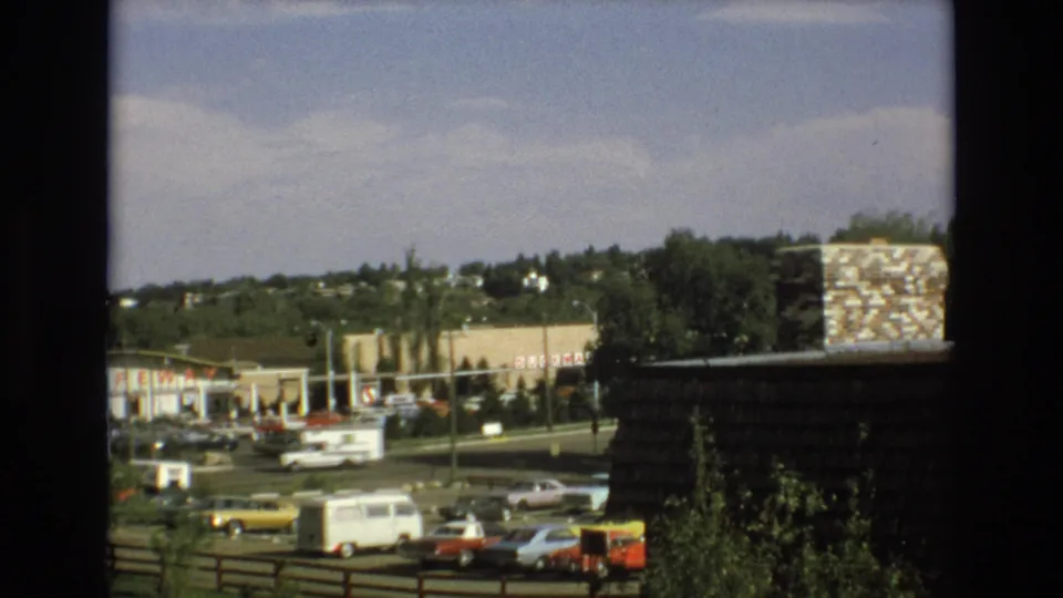 Colorado Springs home-movie frame from 1973 showing a suburban shopping center, Safeway sign, rows of cars, a white van, foreground trees, and window-frame edges.