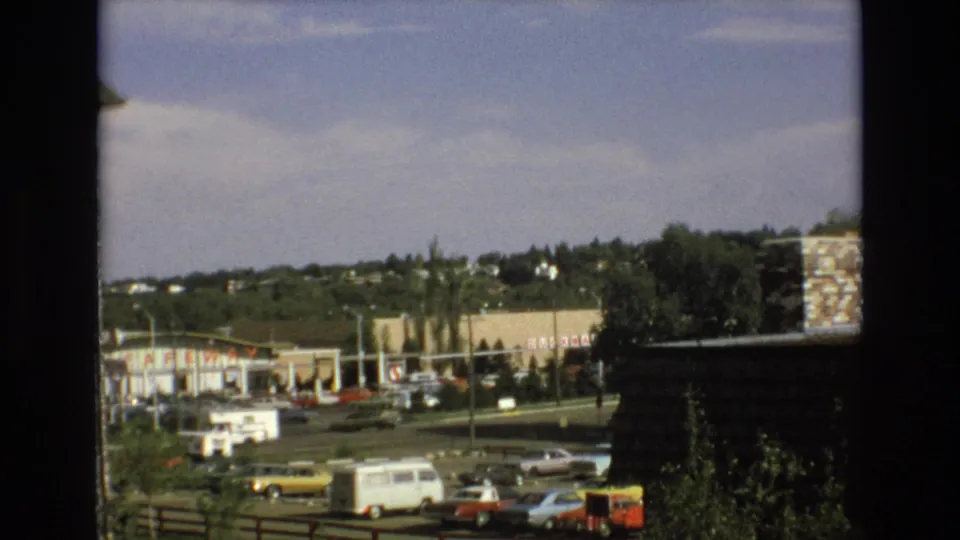 1973 Colorado Springs frame of a suburban commercial area with Safeway signage, a parking lot, classic cars, a white van, roadside poles, trees, and a dark foreground structure.