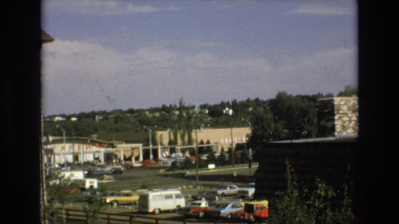 1973 Colorado Springs home-movie view through a dark window or balcony frame toward a roadside commercial area with parked classic cars and distant hills.